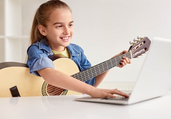 Girl holding an acoustic guitar and watching a tutoril on a laptop computer